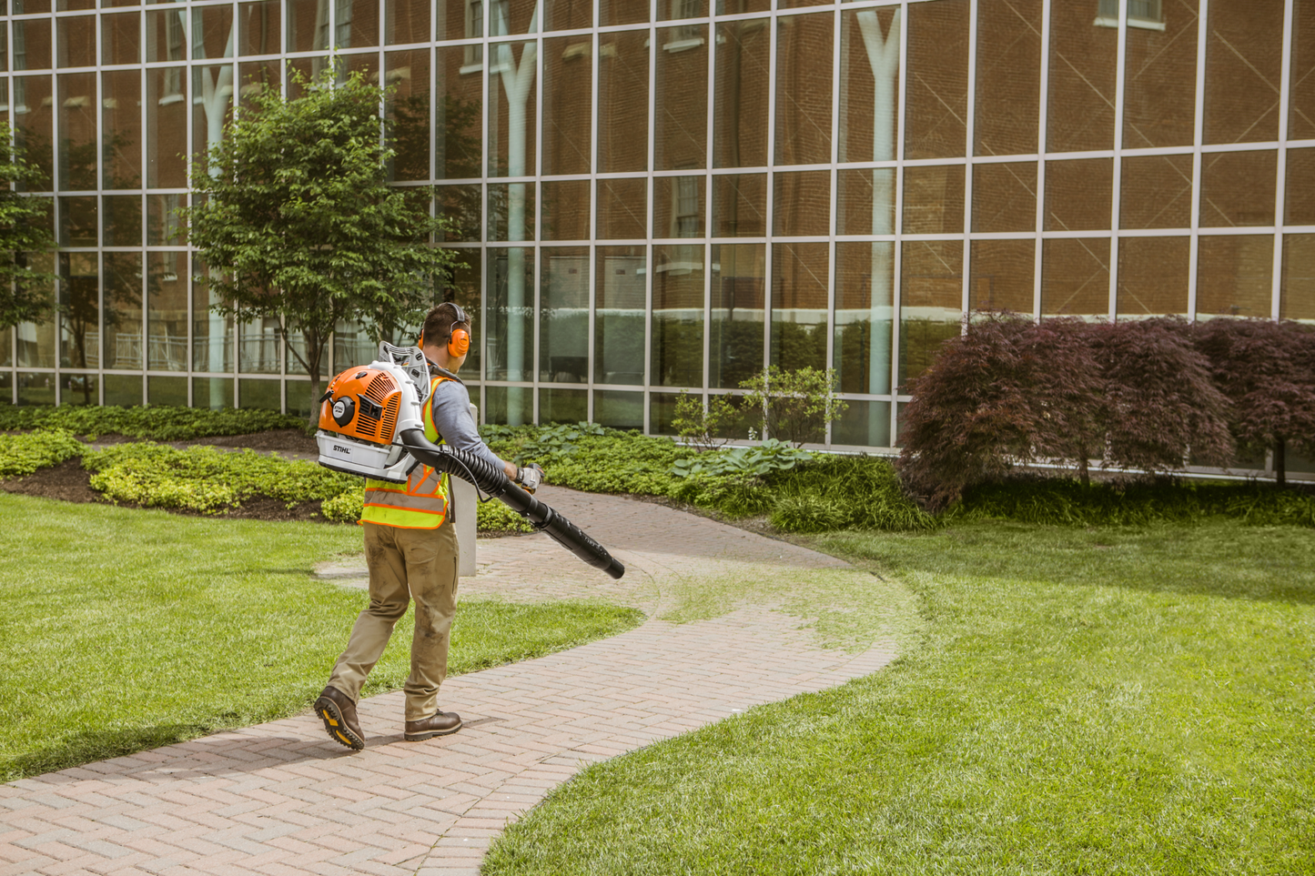 Person using orange and white STIHL backpack blower with protective gear to maintain landscaping near a commercial building