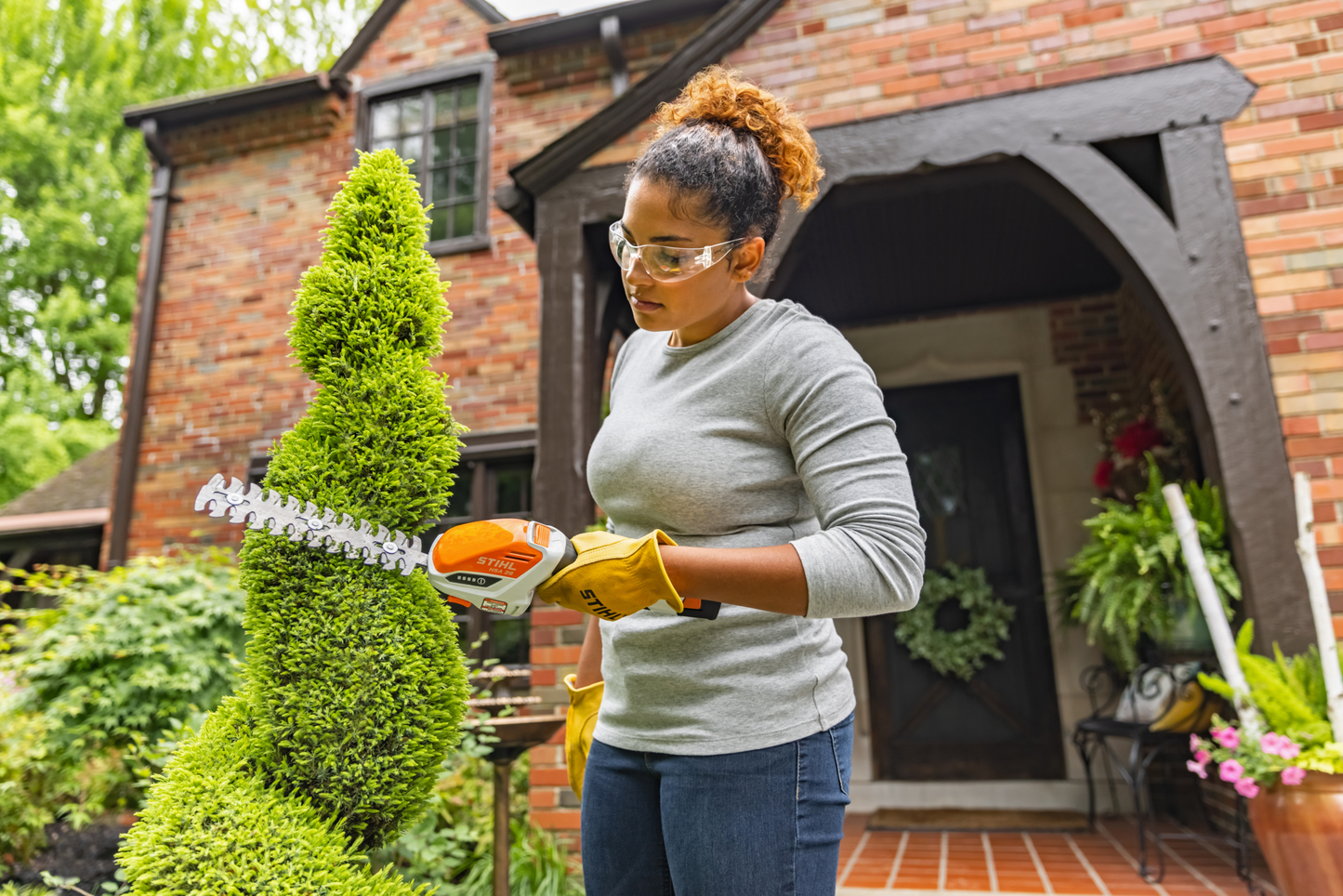Woman wearing safety glasses and gloves shaping spiral topiary with compact orange handheld battery-powered hedge trimmer