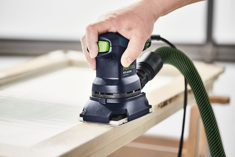 Hand operating a compact black Festool sander with green button and dust extraction on wooden frame for home improvement