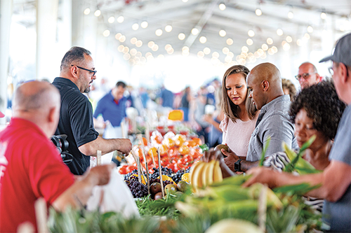 Crowded farmers market scene with shoppers selecting fresh fruits and vegetables under a bright, open-air tent filled with natural light.