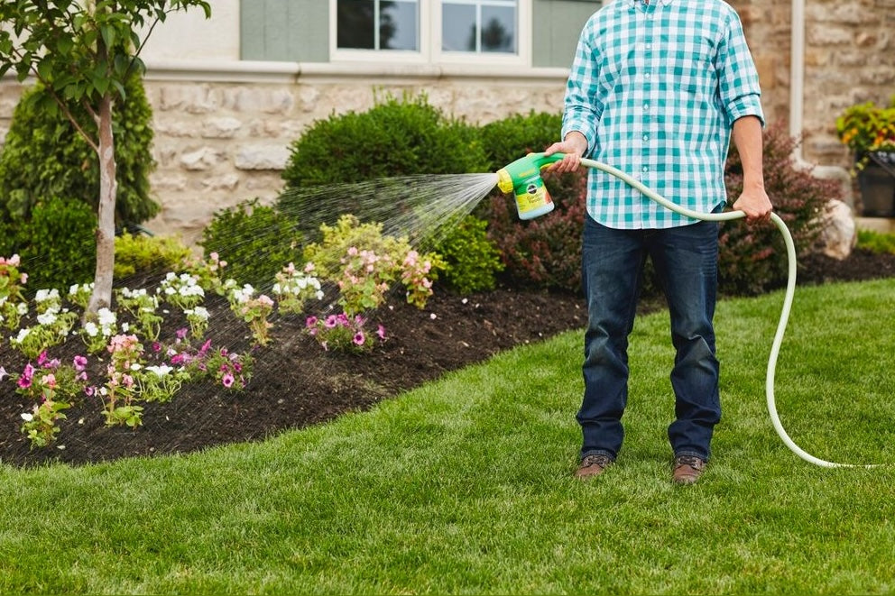 Person using handheld green and yellow garden feeder to water flowers in a landscaped lawn care setting