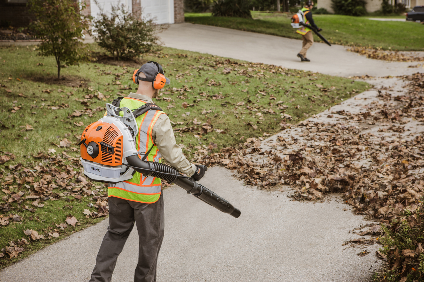 Worker using orange and white backpack blower with protective gear to clear leaves from a suburban driveway