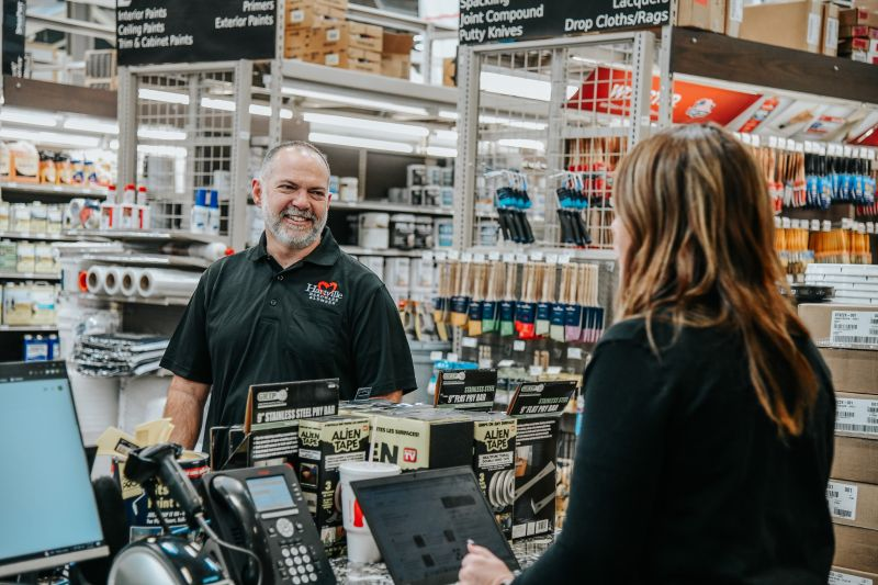 Customer wearing a black jacket speaking with a smiling store employee behind a counter in a hardware store filled with tools and supplies.