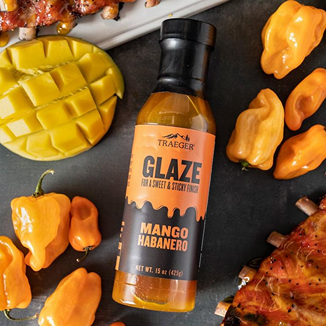 Bottle of Traeger mango habanero glaze surrounded by habanero peppers and sliced mango in outdoor living BBQ sauces collection