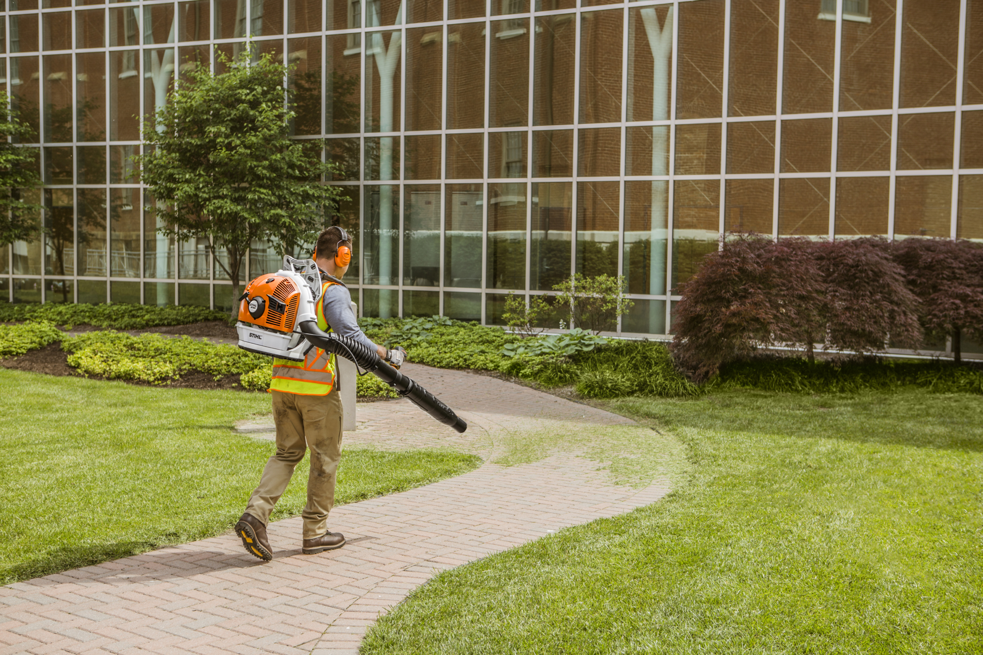 Person using orange and white STIHL backpack blower with protective gear to maintain landscaping near a commercial building