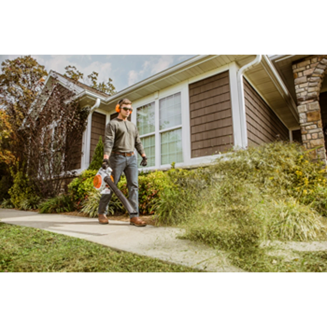 Man using STIHL gas-powered handheld blower to clear debris from a sidewalk outside a home in a garden setting