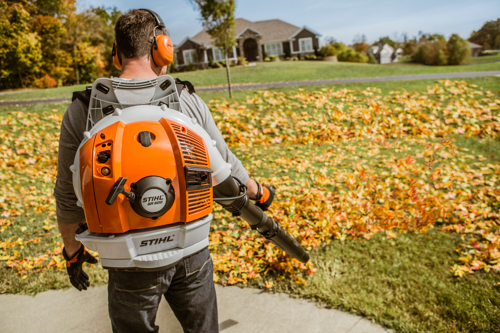 Man using orange and white STIHL backpack blower to clear fallen leaves from lawn in a residential neighborhood