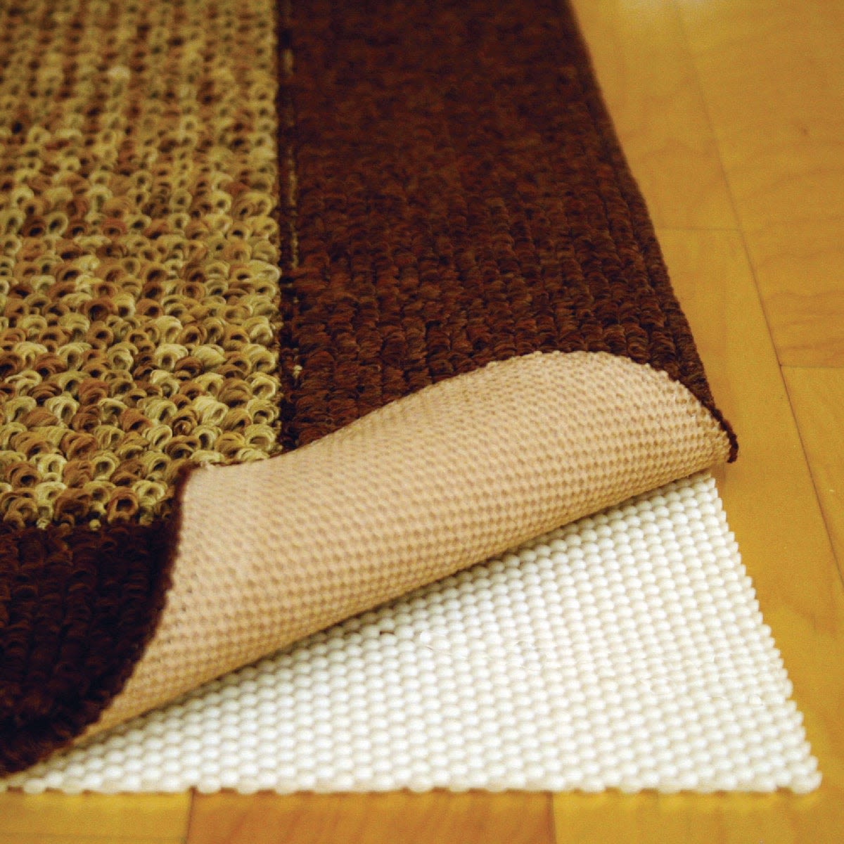 Brown and beige textured area rug corner lifted to show white nonslip rug pad underneath on wood flooring