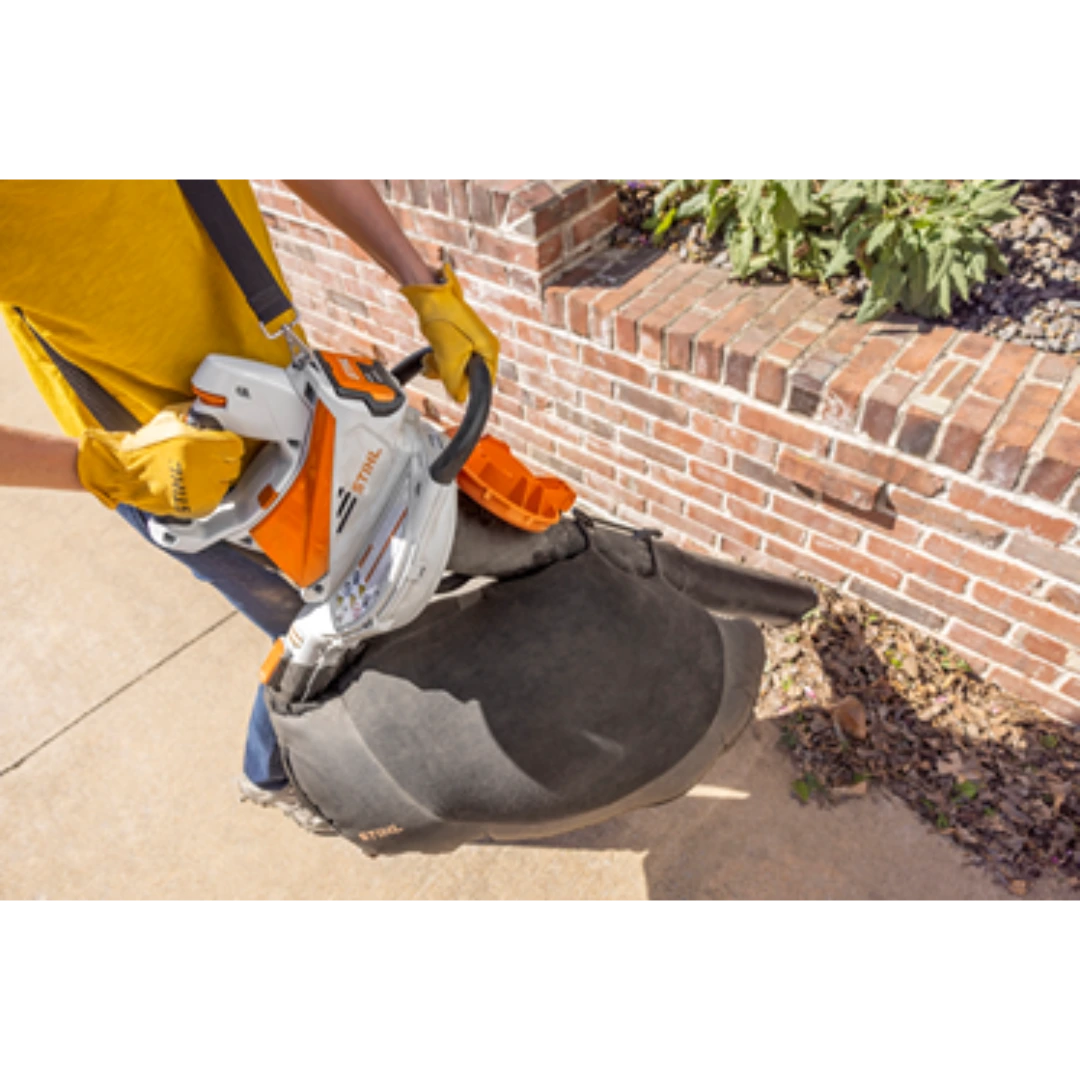 Person wearing yellow gloves operating a white and orange battery-powered garden shredder vacuum with black collection bag outdoors