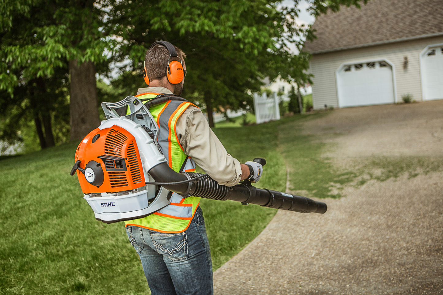 Person wearing protective gear using an orange and white backpack blower for outdoor home maintenance tasks