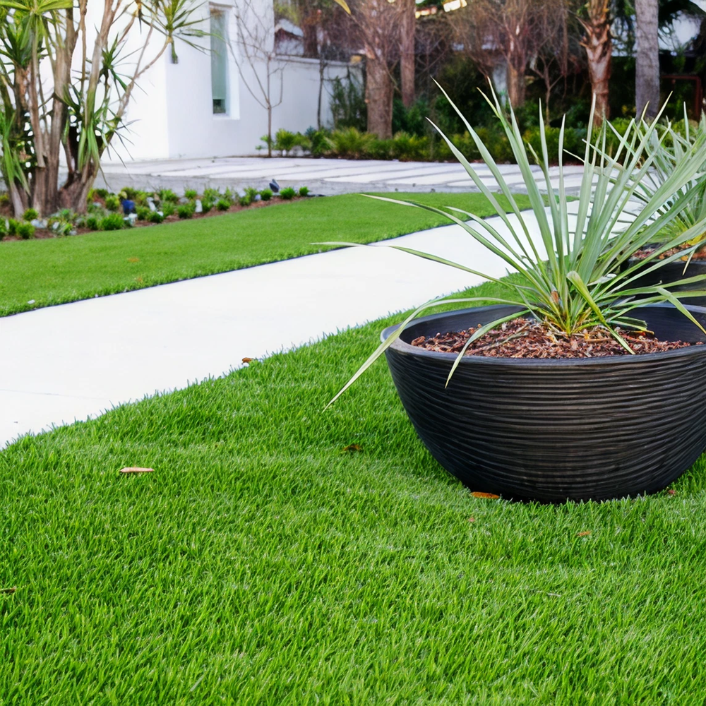 Outdoor scene with a modern white walkway bordered by vibrant green artificial grass and a large black planter holding a spiky green plant near a white building.