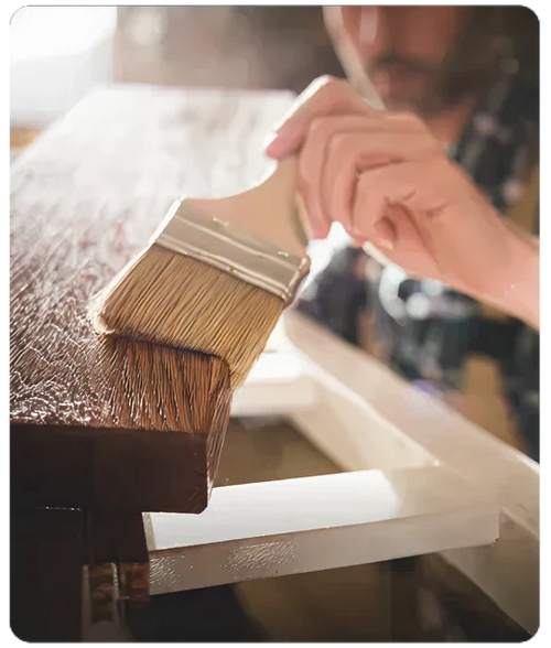 Person applying a coat of varnish on the edge of a wooden surface using a wide paintbrush in a workshop setting