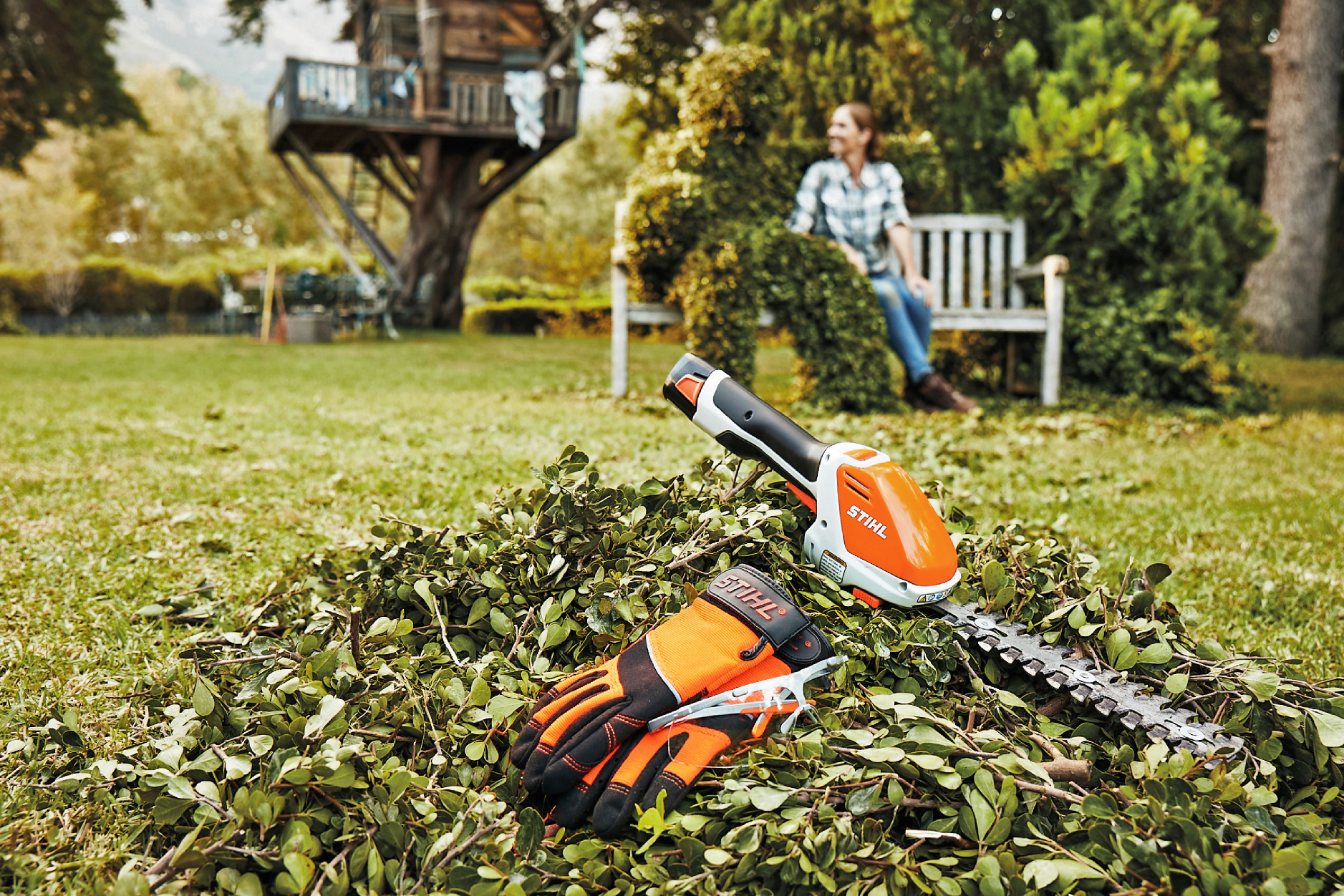 Orange and white cordless handheld hedge trimmer with gloves resting on freshly trimmed green branches outdoors
