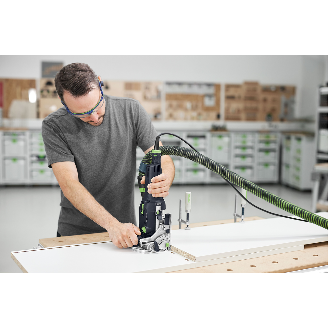 Man using a Festool DOMINO joiner with dust extraction hose to cut precise joints on a white wood board in a woodworking workshop surrounded by power tools and storage cabinets.