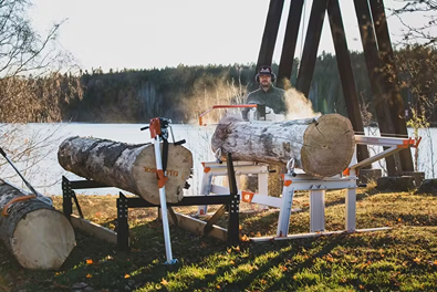 Man using logosol f2+ chain sawmill with two log lifts to cut large logs outdoors near a lake at sunrise or sunset