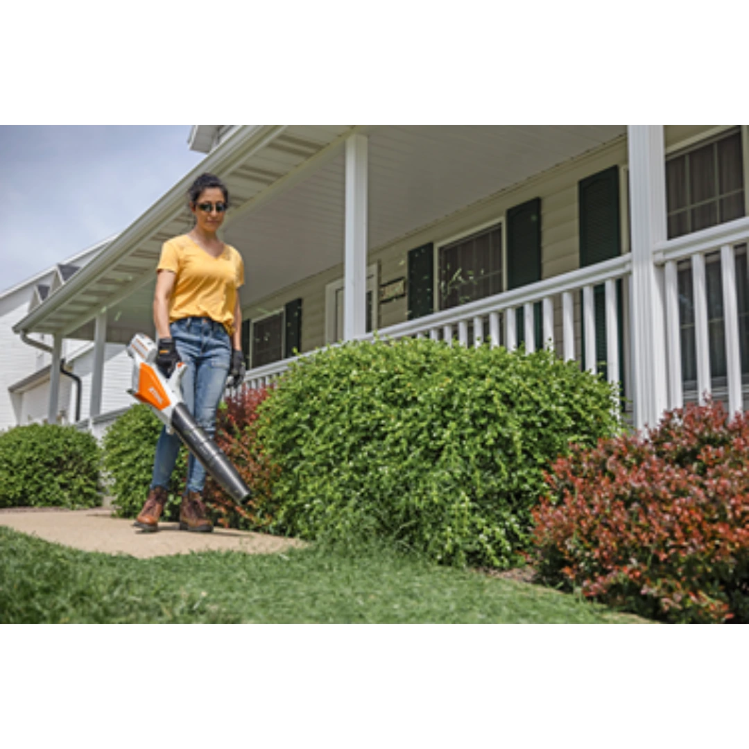 Person using a STIHL cordless battery blower to clear grass clippings on a sidewalk beside bushes at a home