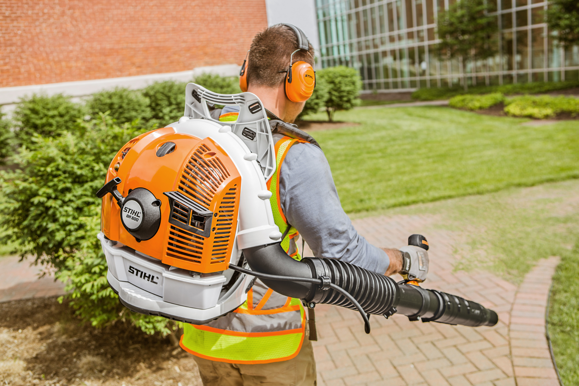 Man wearing ear protection and safety vest using orange and white backpack blower for outdoor landscaping tasks