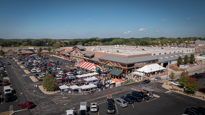 Aerial view of a large retail complex surrounded by busy parking lots, with tents and booths set up near the main entrance under a clear blue sky.