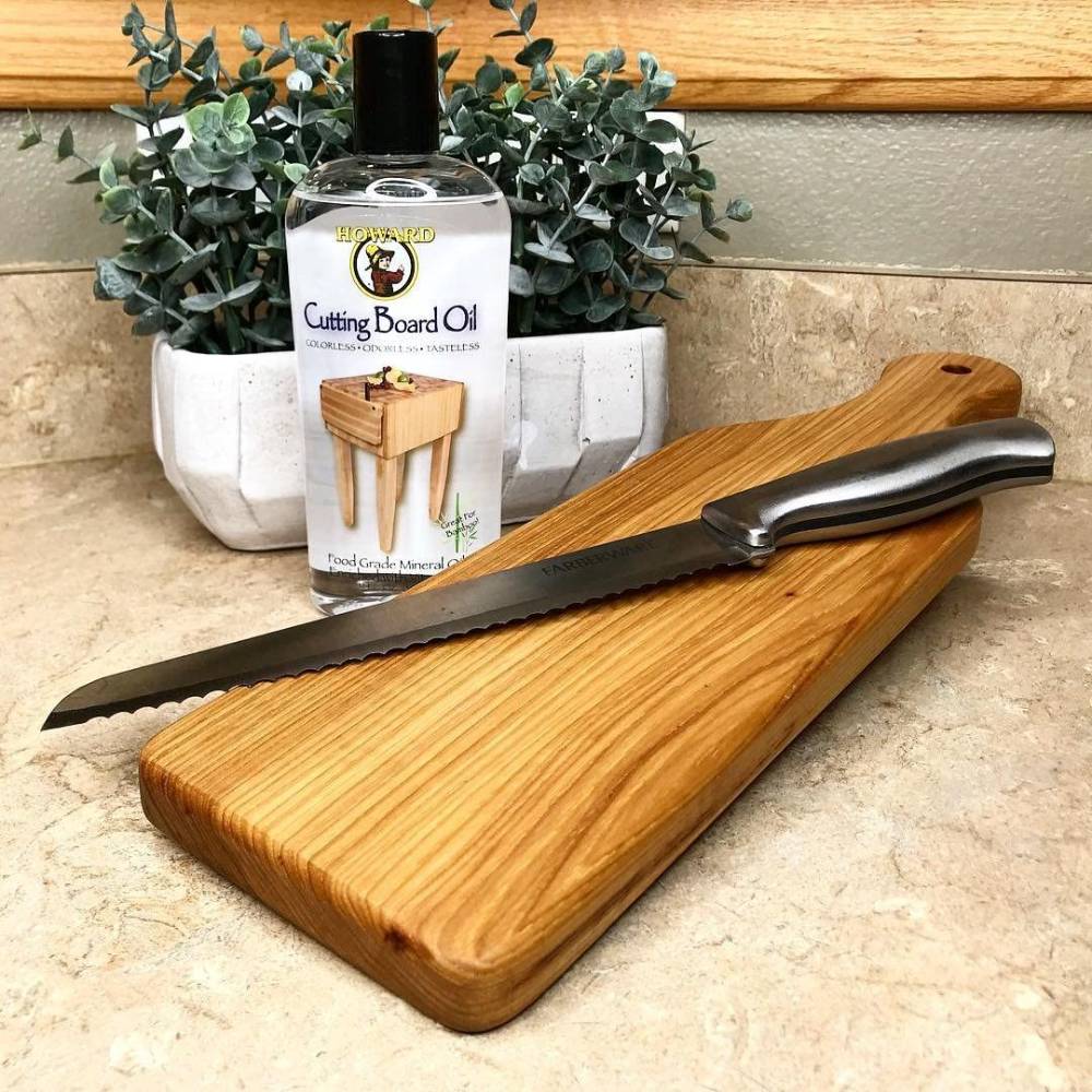 Clear bottle of Howard Cutting Board Oil on countertop next to wooden cutting board and serrated kitchen knife
