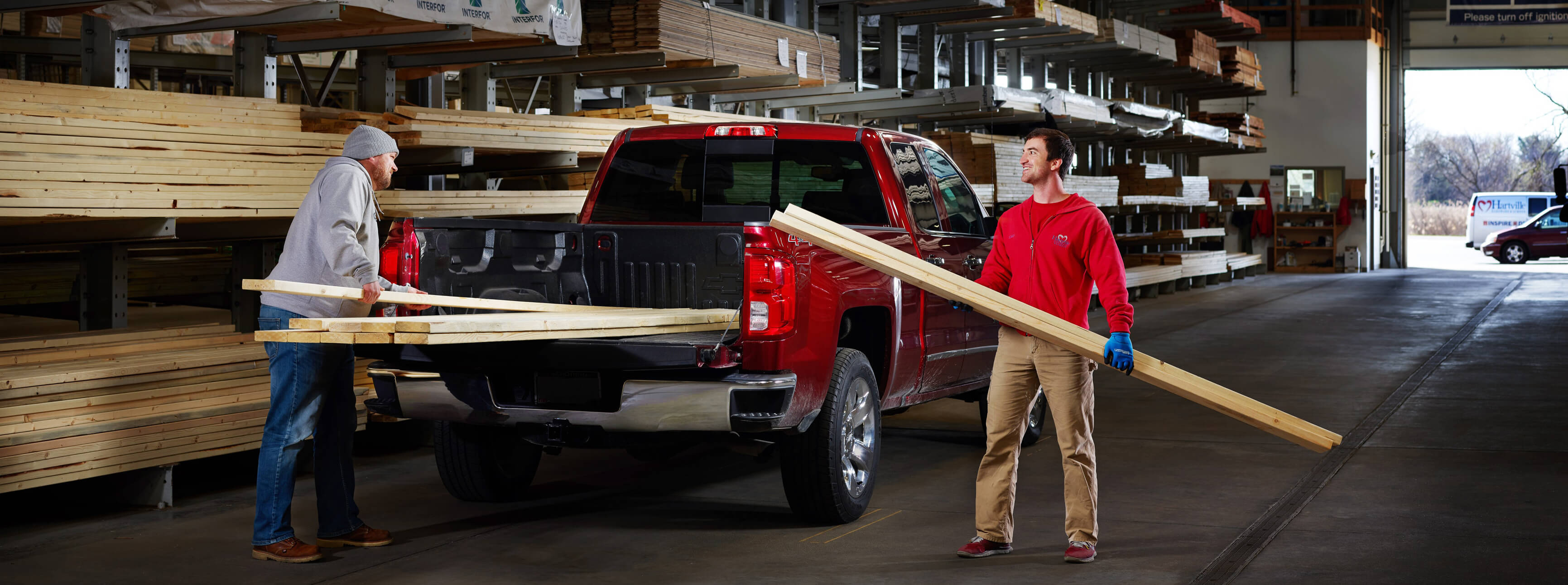 Two men unloading long wooden boards from the back of a red pickup truck inside a large lumber warehouse filled with stacked wood shelves.