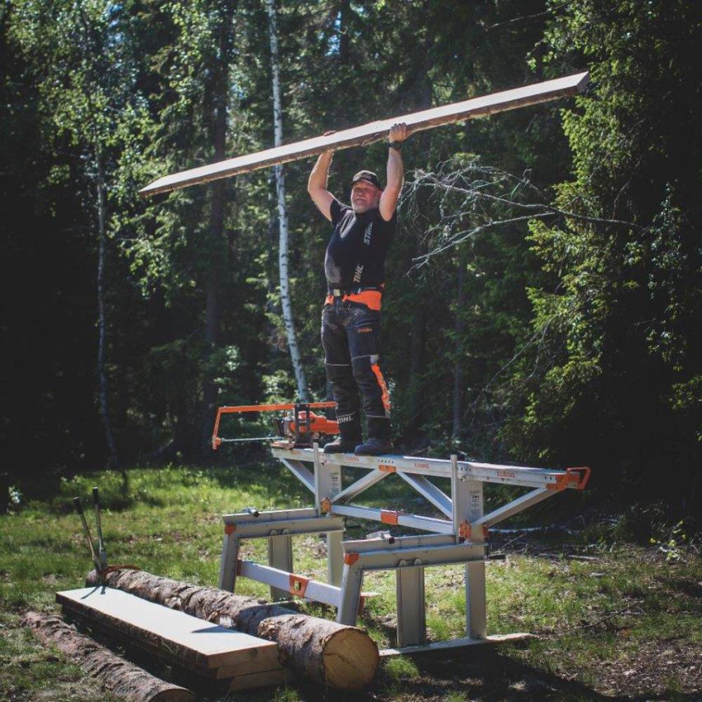 Person holding wooden plank standing on a metallic gray and orange sawmill frame for outdoor woodworking tasks
