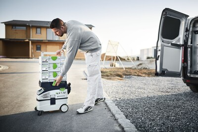 Worker stacking modular gray and green storage boxes on wheeled cart beside open van for jobsite tool storage