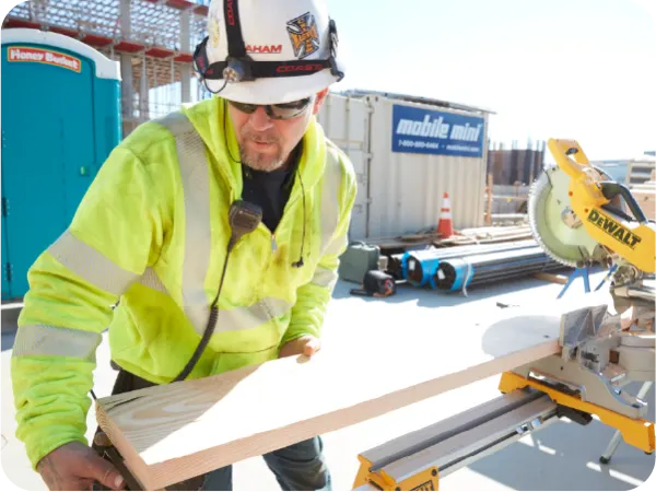 Construction worker wearing high-visibility yellow jacket and hard hat measures a wooden board near a miter saw on an outdoor job site.