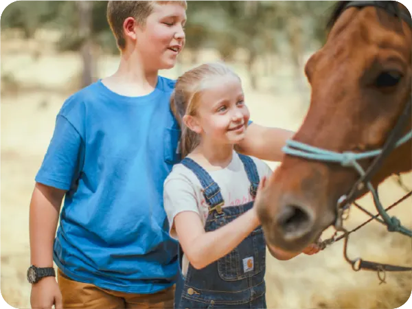 Young boy and girl smiling while petting a brown horse outdoors in a sunny, natural setting
