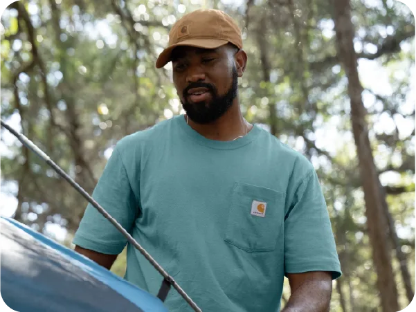 Man in a brown cap and teal shirt inspecting or working on a vehicle outdoors with blurred trees in the background.