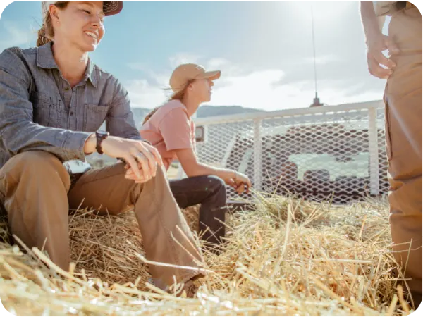 Three people wearing casual outdoor clothing sitting and standing on a bed of hay in a sunlit rural setting with a metal fence in the background.