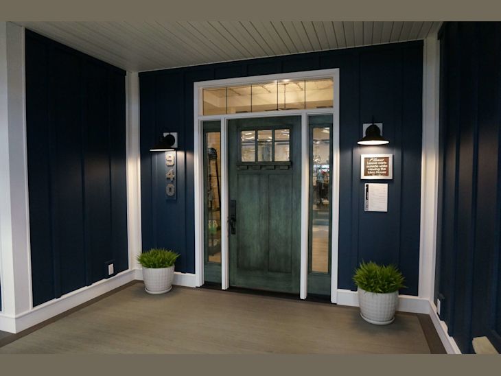 Covered porch with dark blue vertical siding and a green front door illuminated by black wall sconces, flanked by two white plant pots with greenery