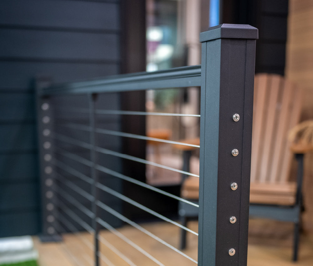 Close-up view of a modern dark gray metal railing with horizontal cables on a deck, with blurred outdoor furniture and siding in the background.