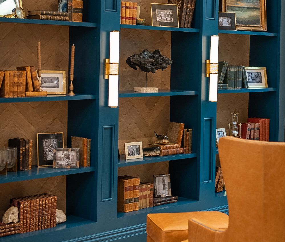 Built-in blue shelving unit filled with vintage books, framed black-and-white photos, decorative objects, and illuminated by vertical wall sconces beside a tan leather chair.