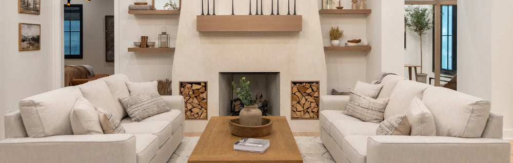 Spacious living area with matching light beige sofas facing a central wooden coffee table, flanked by a stone fireplace with built-in wood storage and minimalist shelving.