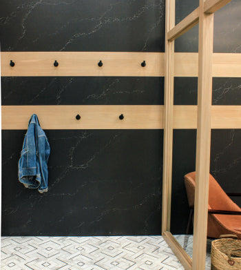 Minimalist entryway with matte black marble-effect wall panels featuring horizontal wooden slats with black hooks, one holding a denim jacket, next to a glass partition and a woven basket on patterned floor tiles.