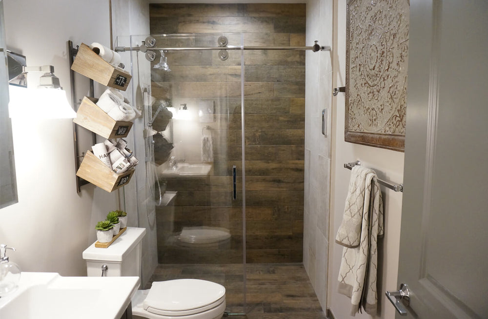 Compact bathroom with glass sliding shower doors, wood-look tile walls, white toilet, angled wall shelves holding towels, and neutral decor.
