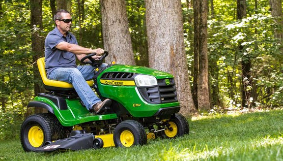 Man wearing sunglasses and a gray shirt riding a green and yellow lawn tractor mowing grass near tall trees in a wooded yard