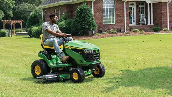 Man riding a green and yellow lawn tractor mowing a well-kept lawn in front of a brick house with a porch and landscaped garden