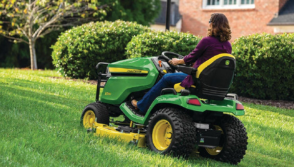 Person wearing a purple top operating a green and yellow riding lawn mower on a lush green lawn in a residential garden with bushes and a brick house nearby