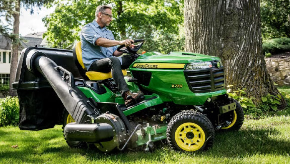 Man wearing glasses and a blue shirt operating a green and yellow riding lawn mower on a shaded grassy area near large trees in a residential yard