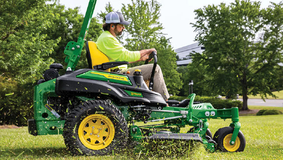 Man wearing a cap and bright yellow shirt driving a green and yellow zero-turn mower on a sunny lawn surrounded by trees and shrubs