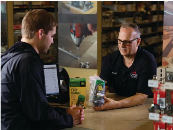 A store employee smiling and assisting a customer across a counter, with engine maintenance items and a computer visible in the background.