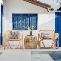 Outdoor seating area with two natural rattan chairs featuring patterned cushions, and a matching woven side table topped with a tropical floral arrangement, set against a white wall with blue window shutters.