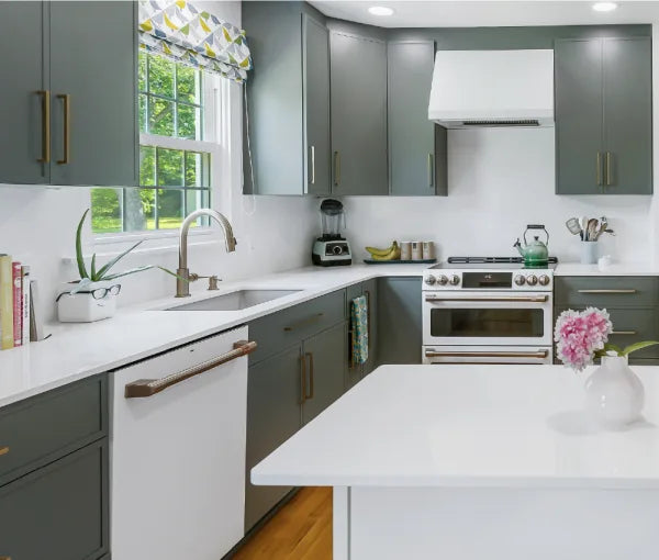 Kitchen with gray cabinetry, white countertops, stainless steel stove, white dishwasher, brass faucet, large window with patterned valance, and a white island with flowers.