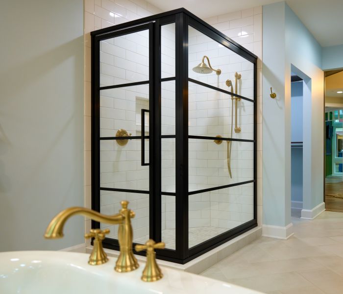 Bathroom featuring a glass shower enclosure with black metal framing, white subway tile walls, gold shower fixtures, and a white sink with gold faucet in the foreground.