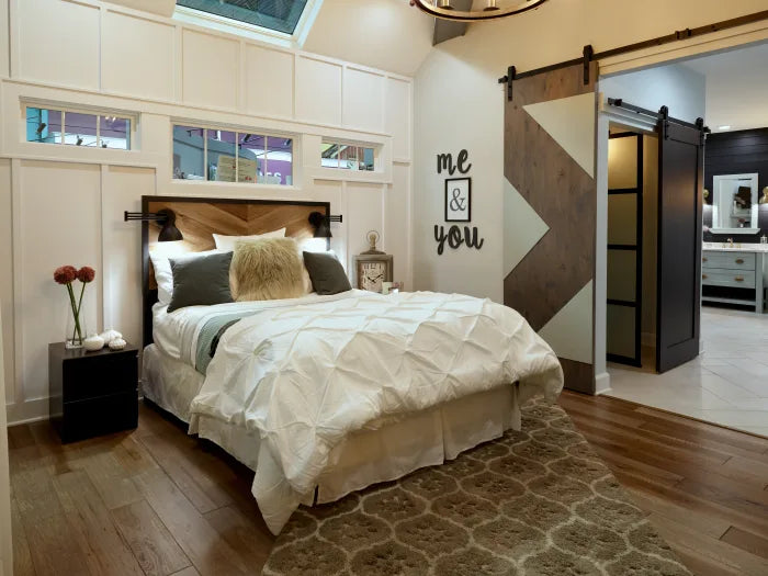 Bedroom with a white textured comforter on a bed featuring a wooden headboard, black wall lamps, a patterned area rug, and sliding barn doors leading to a bathroom.