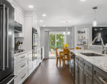 Spacious kitchen with white upper cabinets, gray island with marble countertop, stainless steel appliances, wooden dining table, and sliding glass doors.