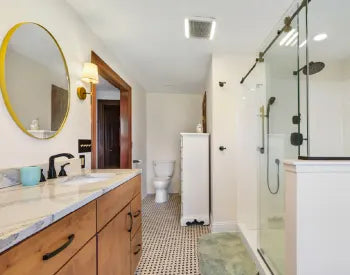 Bright bathroom with wood vanity topped by marble countertop, round gold-framed mirror, black fixtures, glass shower enclosure with black hardware, and patterned tile floor.