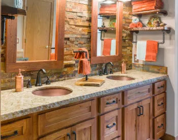Rustic bathroom vanity with dual copper sinks, granite countertop, oil-rubbed bronze faucets, wooden cabinets, and stone accent wall backing two wooden framed mirrors.