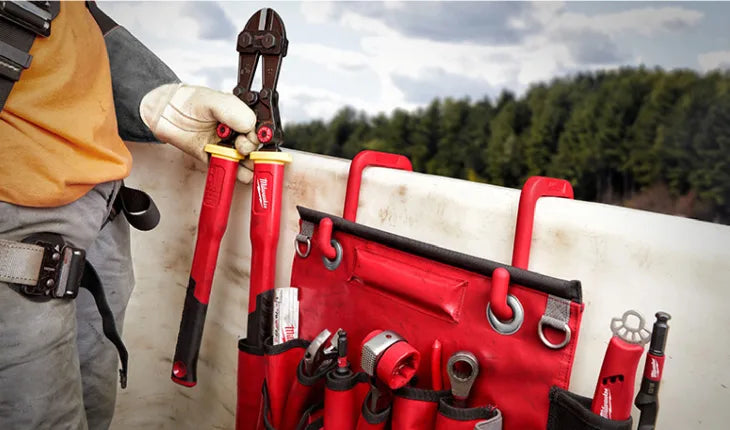 Close-up of a person wearing gloves and work gear holding red-handled bolt cutters next to a red tool pouch attached to a white surface, with a blurred natural background.